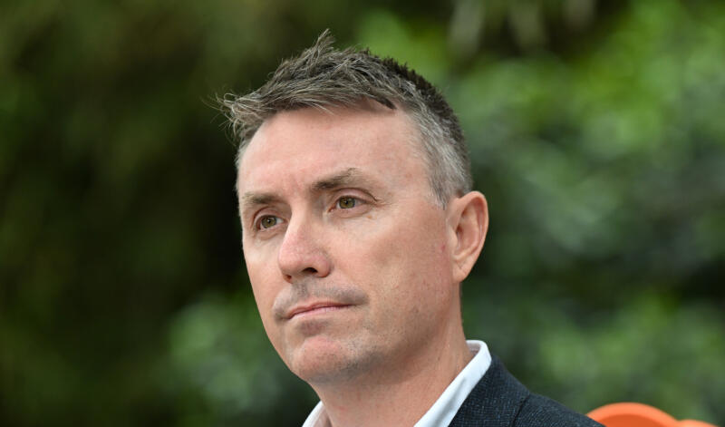 a close-up image of a caucasian man looking serious with green shrubbery in the background