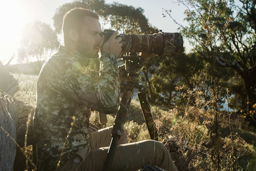 Vaughan King sits in camouflage at a tripod looking through a camera with a long lens.