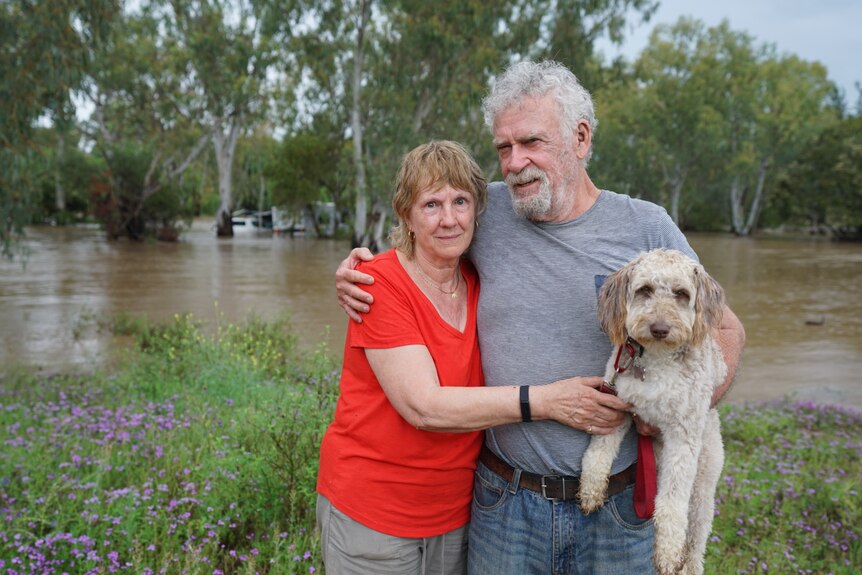 Jan and John and their dog hug for a photo in front of a swollen river. 