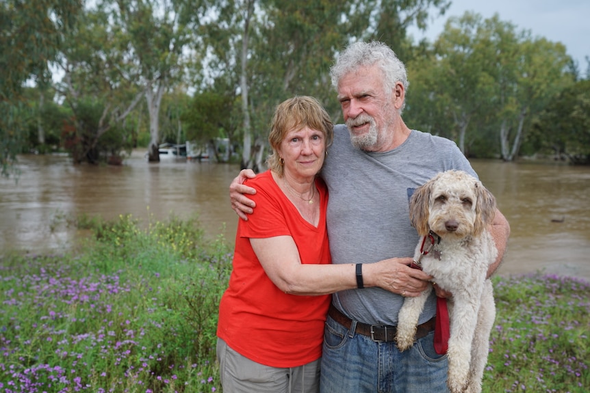 Jan and John and their dog hug for a photo in front of a swollen river. 