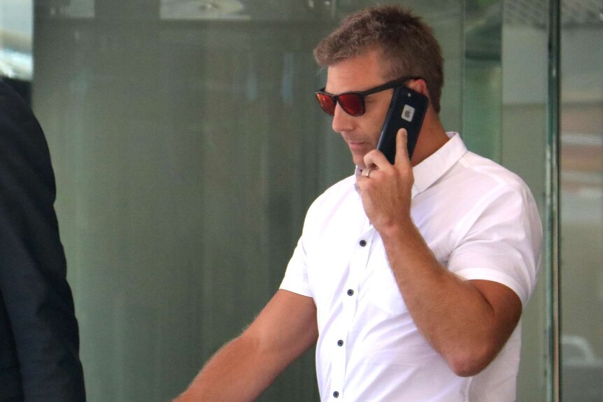 A mid shot of a man in a white shirt and sunglasses talking on a mobile phone as he walks out of a building.
