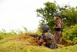 Two military guards hold guns behind a mud embankment. One is standing, one is crouching.