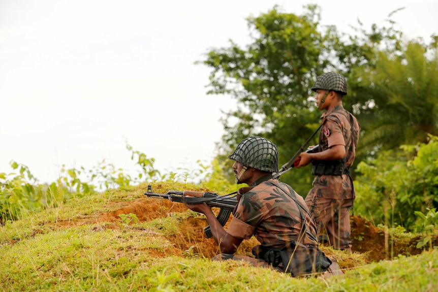 Two military guards hold guns behind a mud embankment. One is standing, one is crouching.