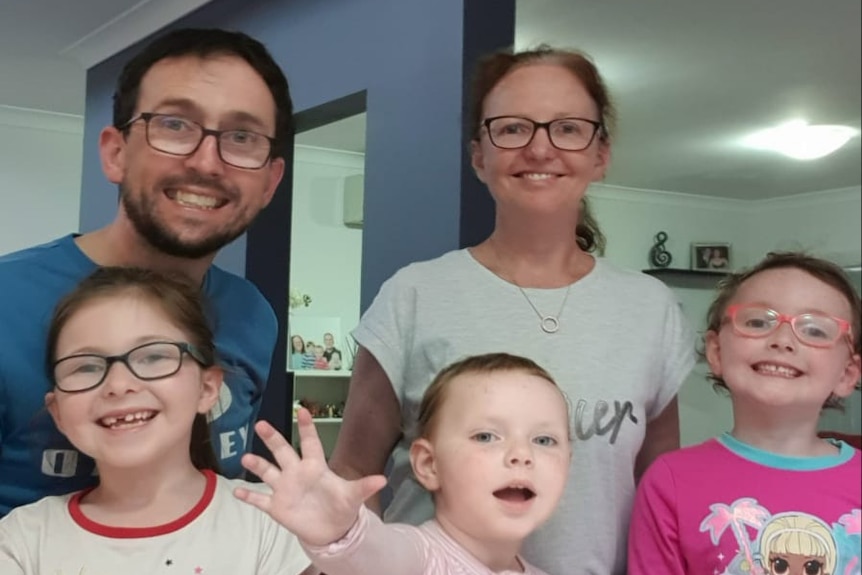 A man and woman wearing glasses stand behind their three young daughters who are smiling at the camera.