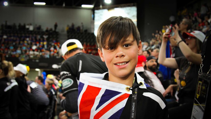 A young boy smiles at the camera whilst draped in a New Zealand flag.