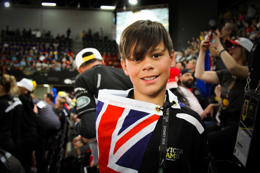 A young boy smiles at the camera whilst draped in a New Zealand flag.