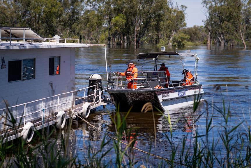 A boat with SES signage and three people wearing orange jumpsuits alongside a houseboat on the river