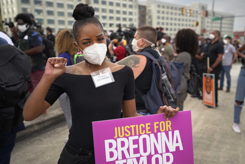 A black woman wearing black clothes and a white mask holds a sign saying justice for Breonna Taylor with people crowded behind