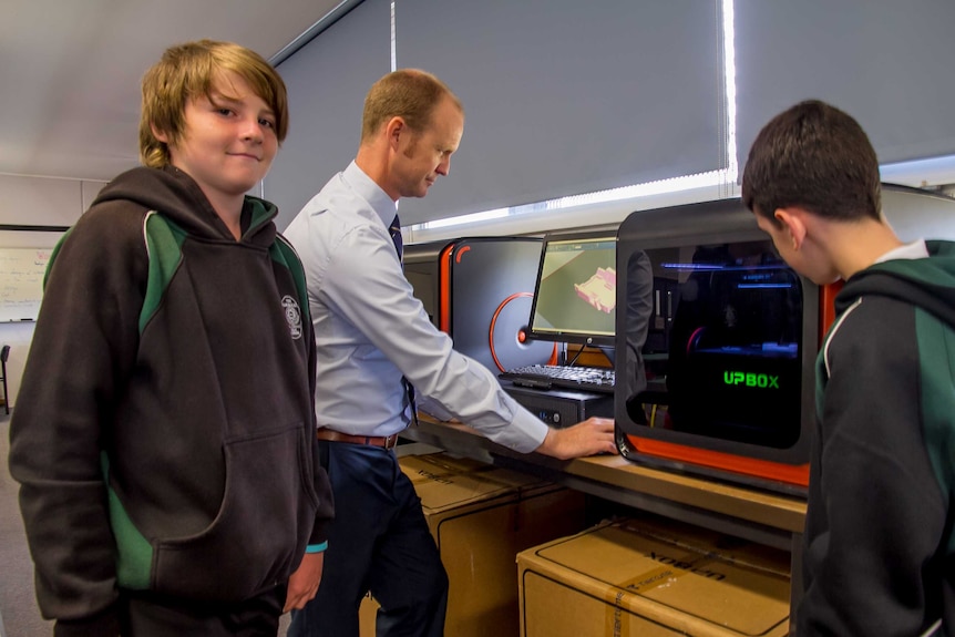 Two boys in school uniform and am an in a suit looking at screens and big 3d printer boxes