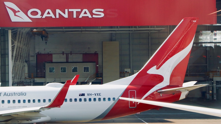 The tail of a Qantas plane in front of a Qantas shed on a tarmac at an airport.