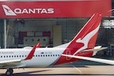 The tail of a Qantas plane in front of a Qantas shed on a tarmac at an airport.