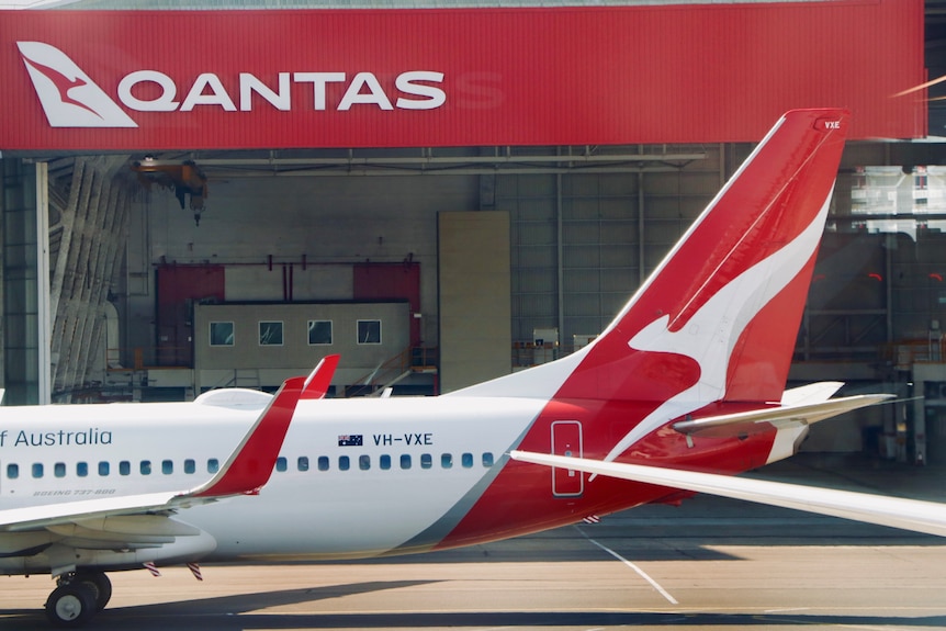 The tail of a Qantas plane in front of a Qantas shed on a tarmac at an airport.