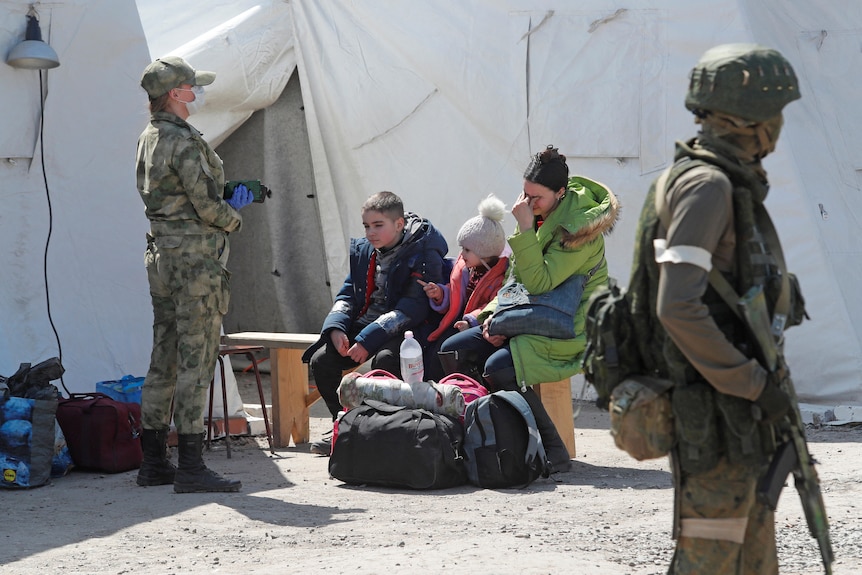 A woman and children sit outside tents as guards stand nearby.