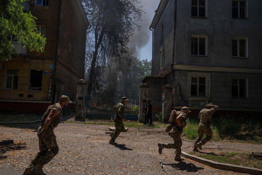 Four men in green uniforms run along a street littered with leaves. Between two buildings, a plume of smoke rises