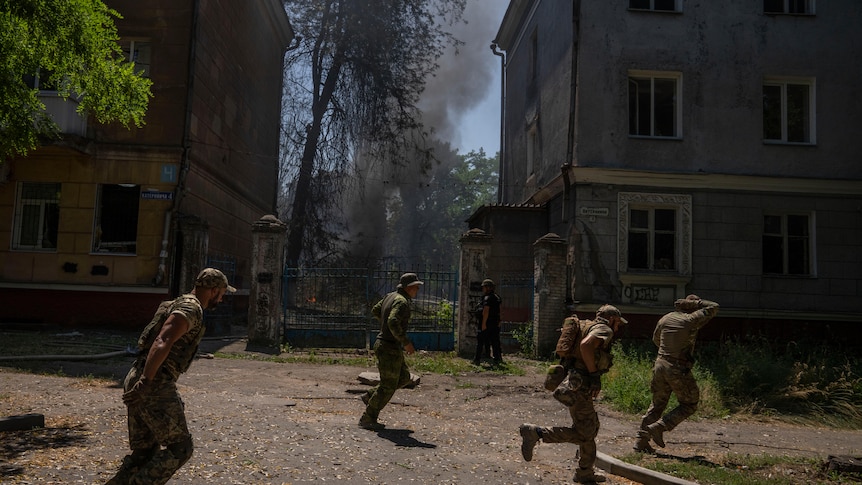 Men in military gear run left to right. Behind them between two buildings smoke rises from a large tree.