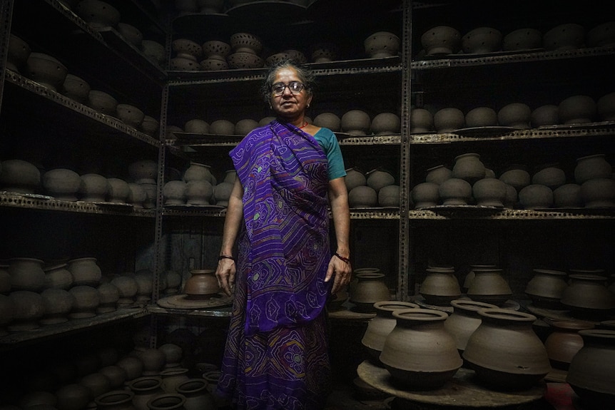 A woman in a sari stands in front of shelves lined with black pots 