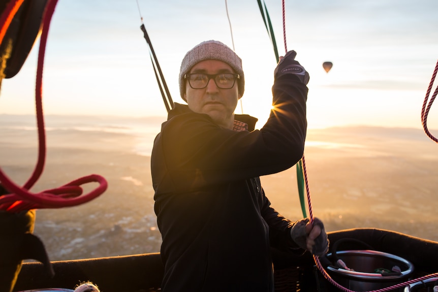 Pilot Chris Shorten pulls on ropes to steer his hot air balloon, warm dawn light behind him.