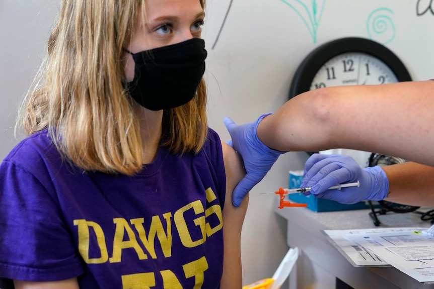 A blond girl in a blue t-shirt with the left sleeve rolled up receives an injection