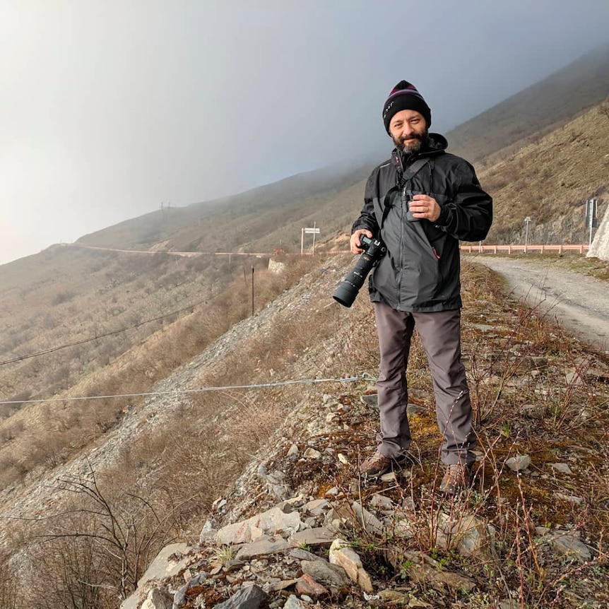 Man standing on a desolate hillside next to a road. He has a beard, he's wearing a black jacket and hat, holding a camera.