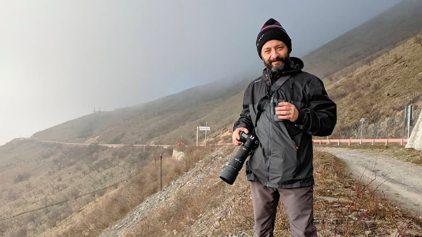 Man standing on a desolate hillside next to a road. He has a beard, he's wearing a black jacket and hat, holding a camera.