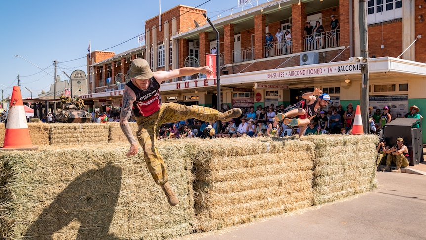 Two men hurdle of row of hay bales in front of an old hotel and hundreds of spectators.