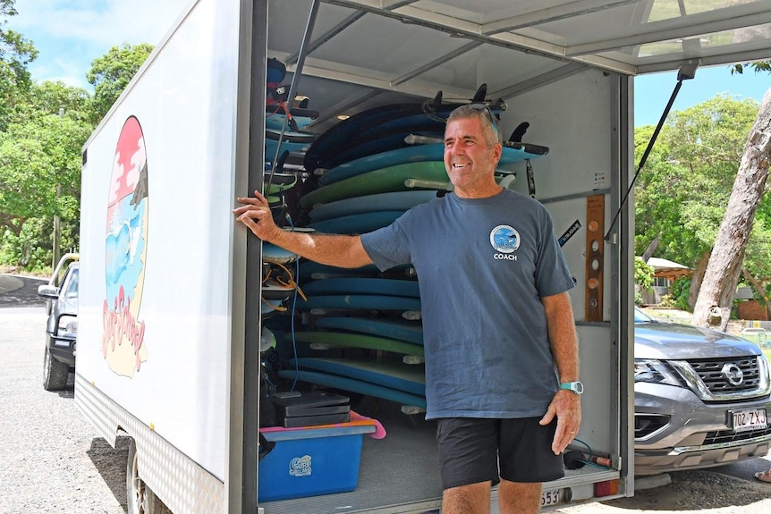 Murray Taylor stands in front of trailer full of surfboards, smiling.