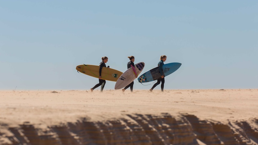 Three women in full wetsuits walk across a windy beach with surfboards