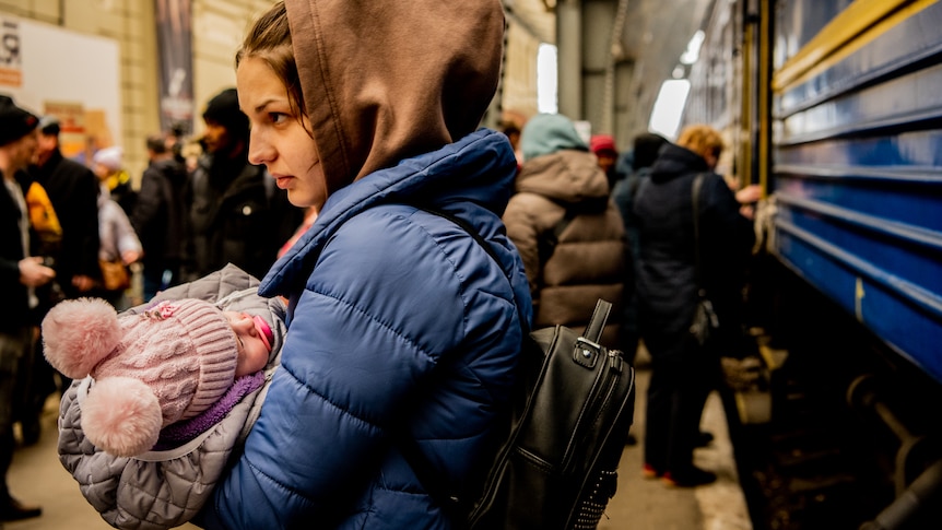 A woman holding a young child stands near a train. Both are wearing winter clothes, and other people stand behind them.