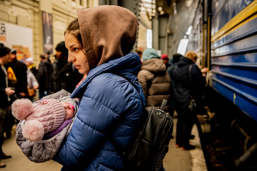 A woman holding a young child stands near a train. Both are wearing winter clothes, and other people stand behind them.