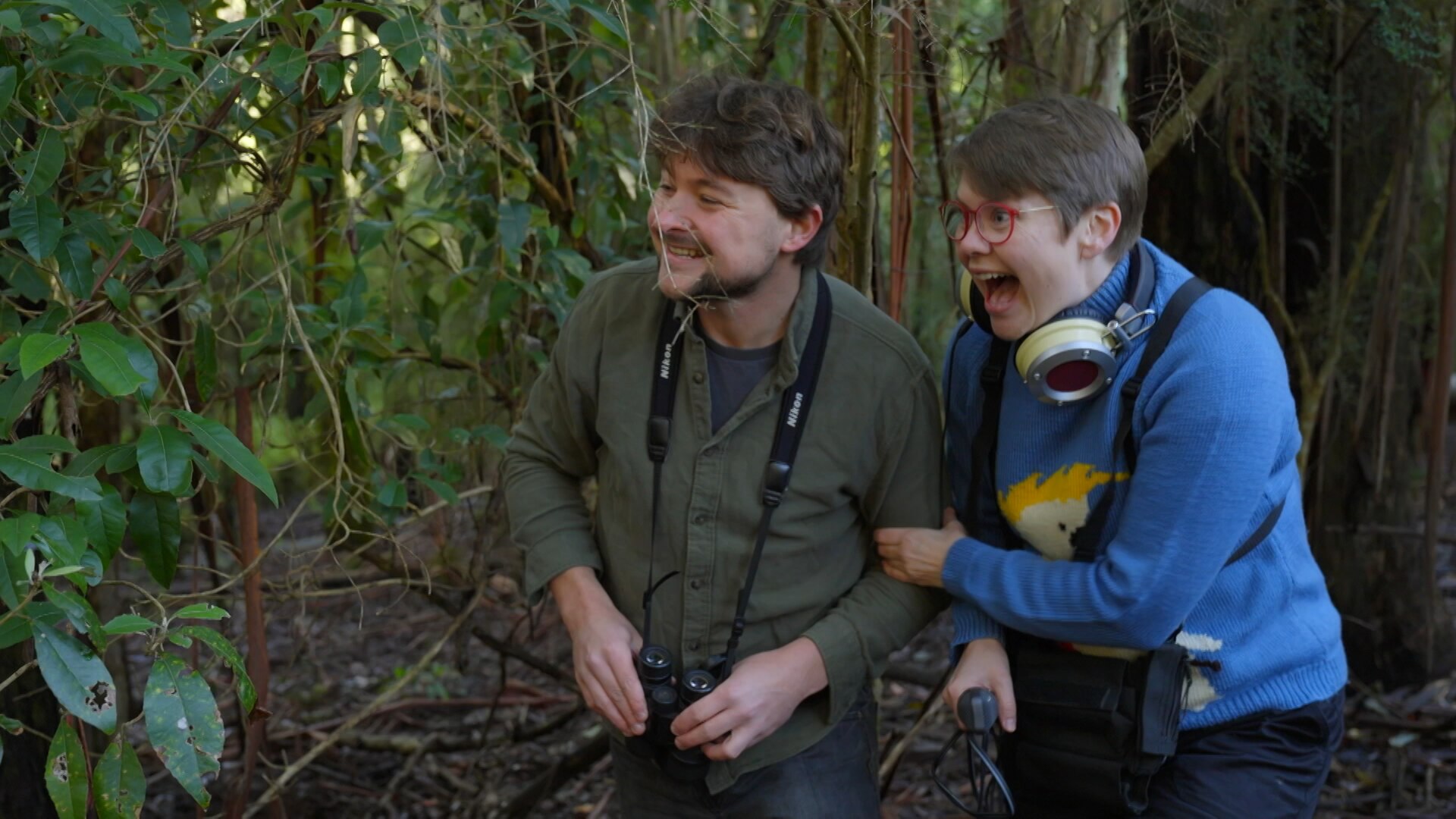Woman wearing headphones with a man with binoculars, standing in a forest with an excited look on faces.