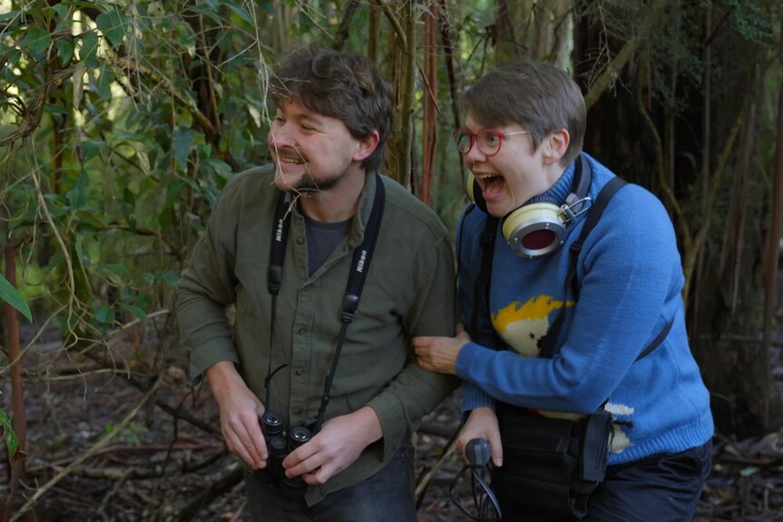 Woman wearing headphones with a man with binoculars, standing in a forest with an excited look on faces.