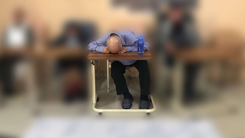 Three men in chairs at an aged care facility, one with his face down on a table.