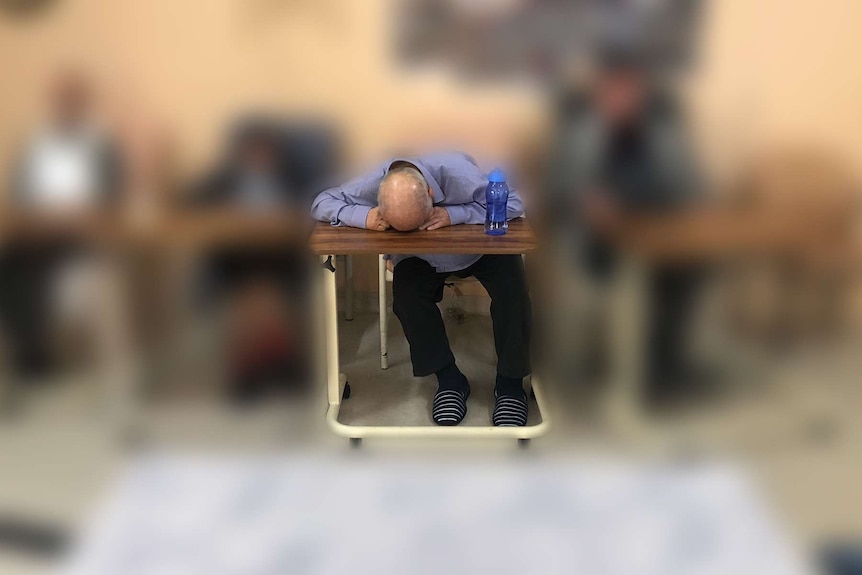 Three men in chairs at an aged care facility, one with his face down on a table.