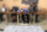 Three men in chairs at an aged care facility, one with his face down on a table.