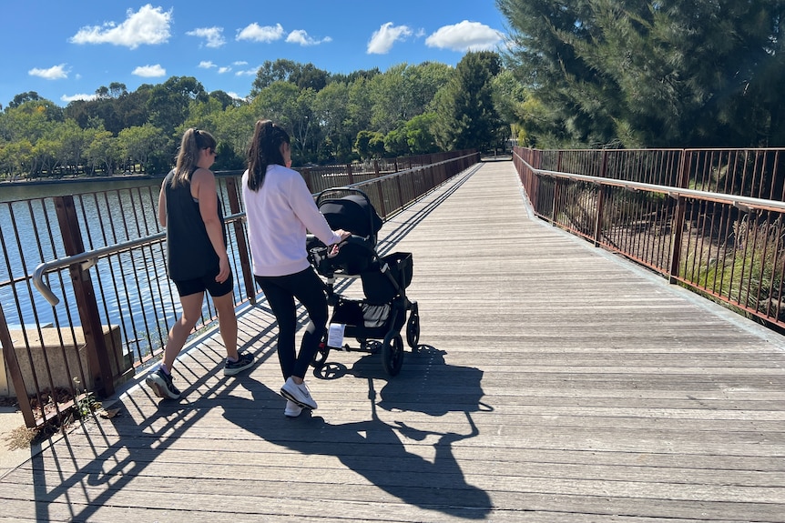 Two women wearing activewear, and one pushing a pram, walk over a bridge over a lake