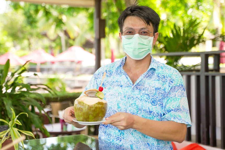 A Thai man in a face mask holds a cocktail served in a coconut 