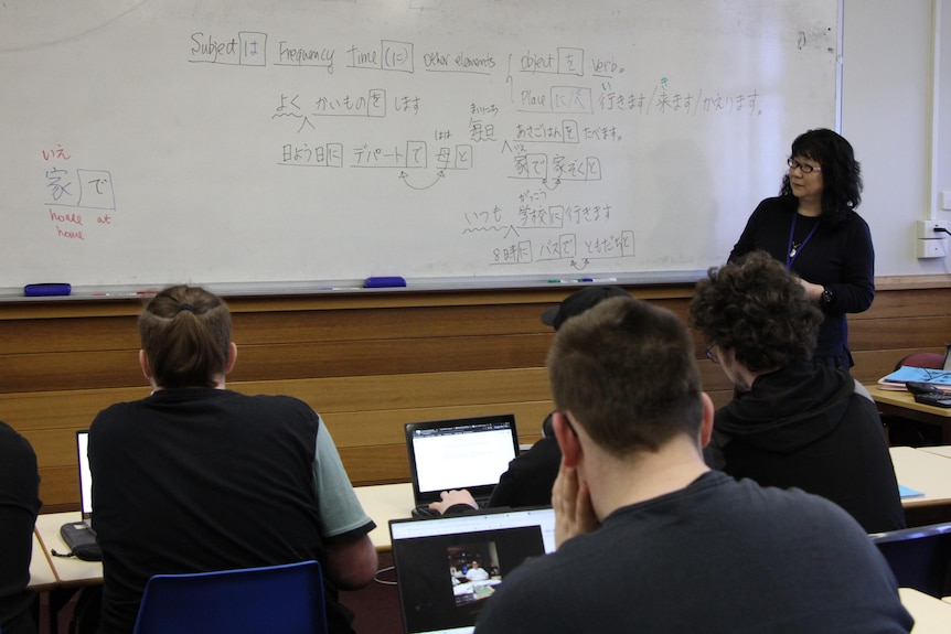 Students look at a whiteboard with a mixture of English and Japanese writing on it, as a teacher stands nearby.