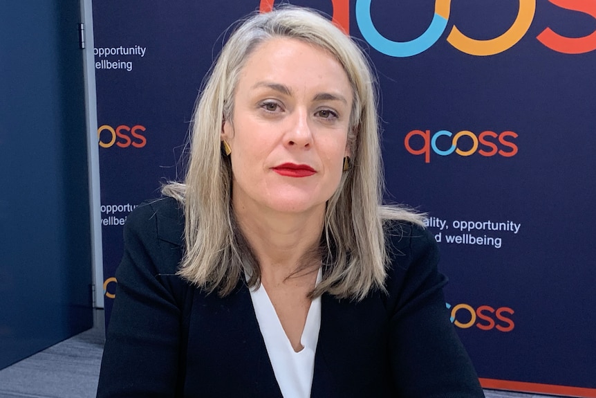 a woman sitting in front of a blue QCOSS banner looking straight at the camera with a serious expression