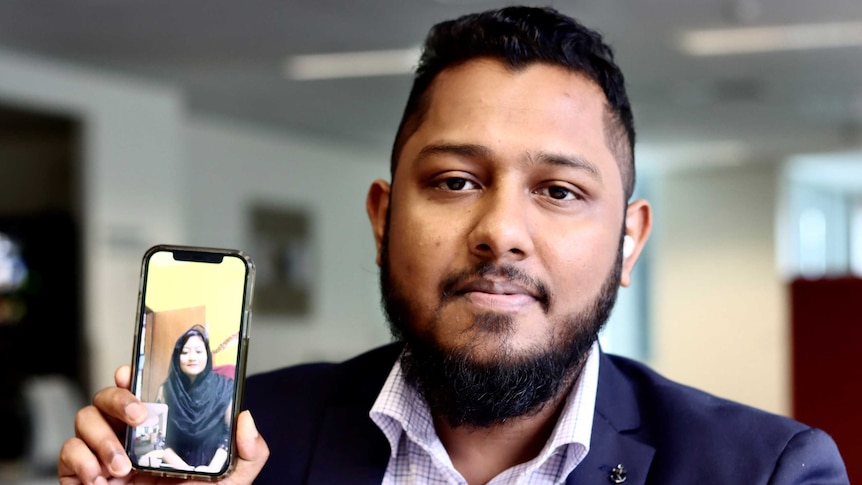 A Bangladeshi man wearing a suit looks inot the camera holding his phone which shows his wife on a video call