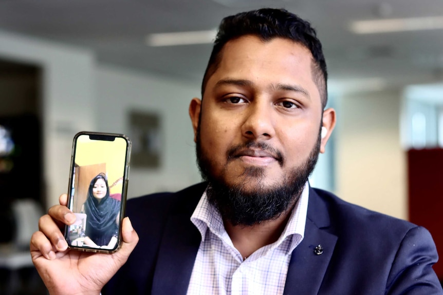 A Bangladeshi man wearing a suit looks inot the camera holding his phone which shows his wife on a video call