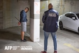 A man stands with his hand cuffed behind his back while a police officer stands watch.