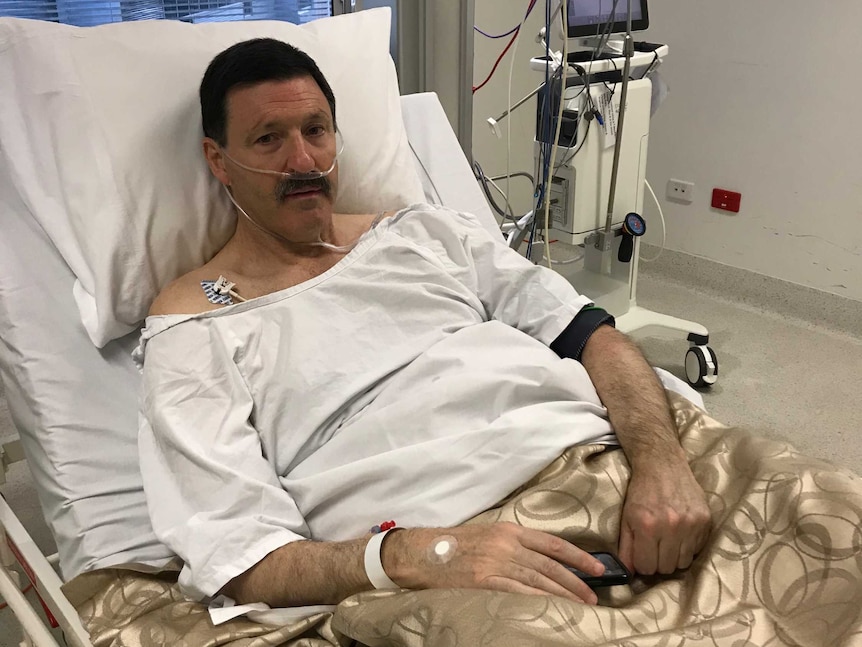 A man in a hospital gown, lying in a hospital bed next to some medical apparatus.