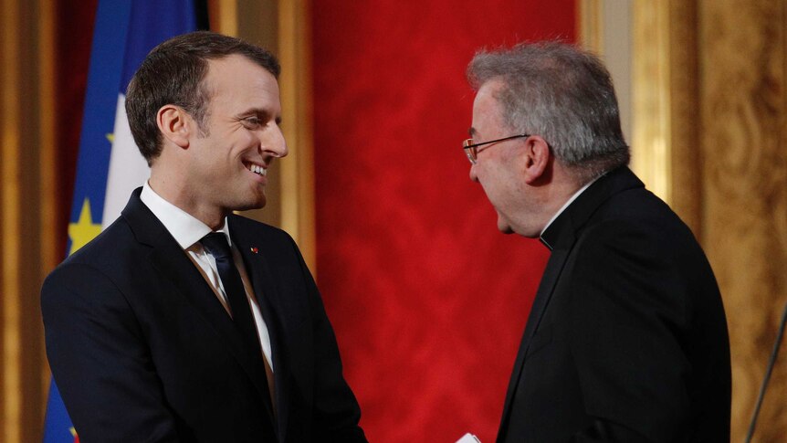 French president Emmanuel Macron is smiling as he speaks to Archbishop Luigi Ventura in front of a flag