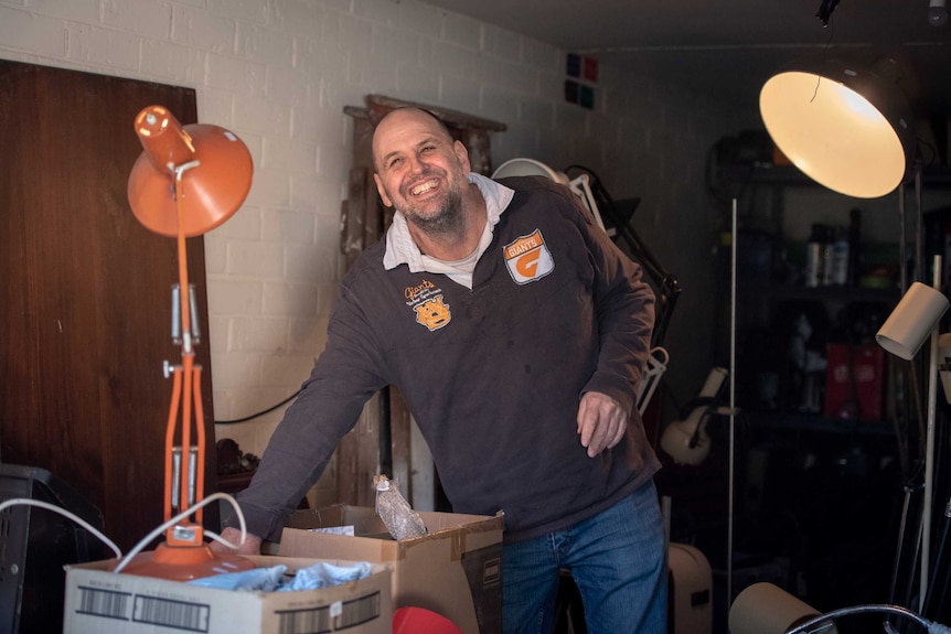 A man stands in a garage, smiling, next to several lights.