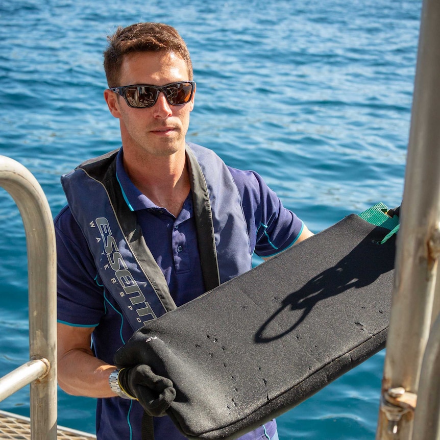A man standing on a boat holds a piece of neoprene fabric, tested for shark bites.