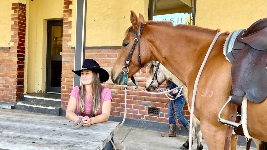 Kylie sitting on a wooden park bench out front of the Toora pub with her brown horse Oaky.