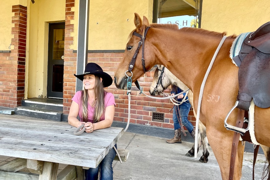 Kylie sitting on a wooden park bench out front of the Toora pub with her brown horse Oaky.