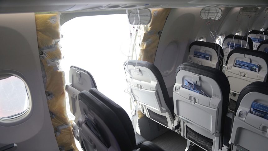 A panel is missing from the side of an aircraft between two rows of passenger seats.