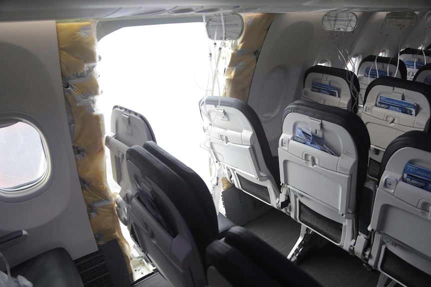 A panel is missing from the side of an aircraft between two rows of passenger seats.