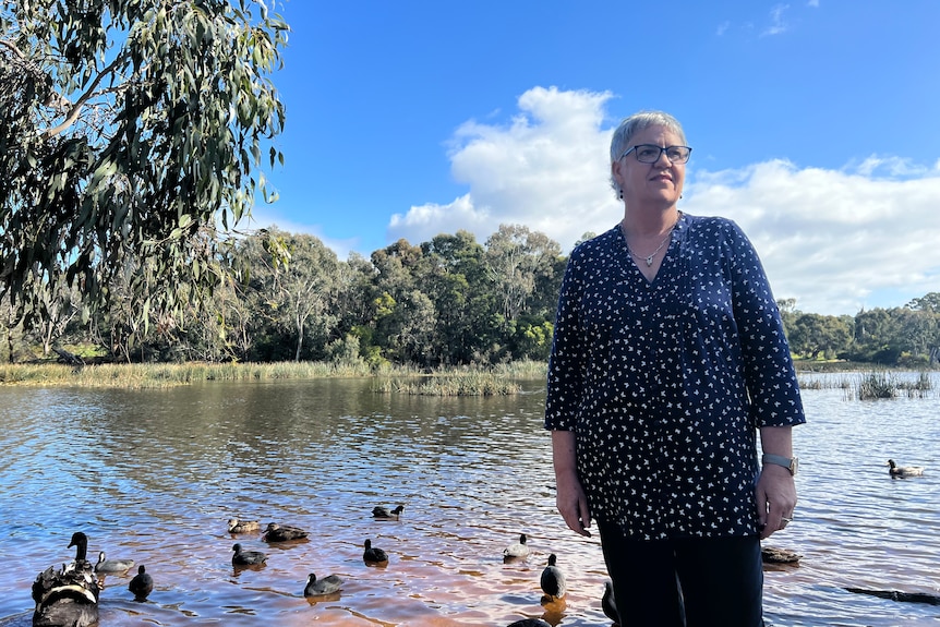 A woman with short hair and spectacles stand in front of a lake with ducks on it.